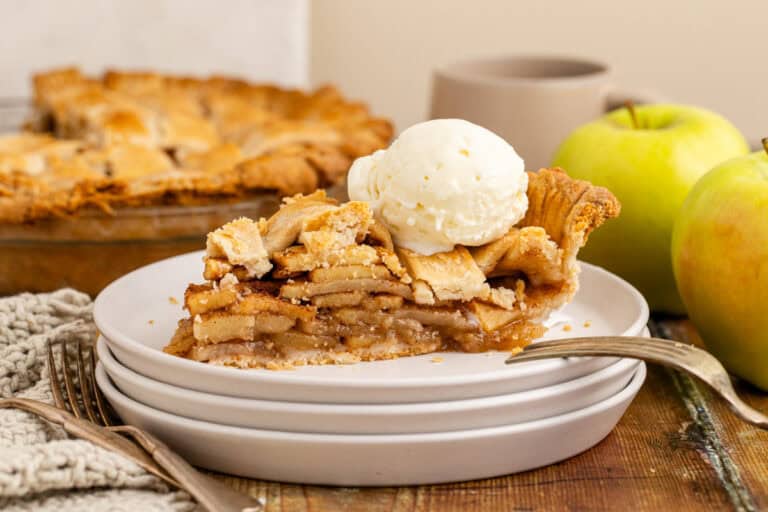 A slice of apple pie topped with vanilla ice cream on a white plate, alongside fresh green apples and a pie in the background.