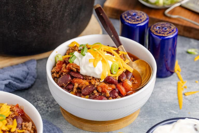 A bowl of chili topped with cheese, sour cream, and green onions sits on a wooden coaster, with a spoon ready for serving.