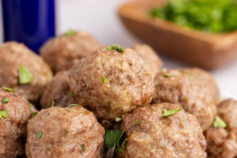 A close-up of golden-brown meatballs garnished with fresh herbs, with a blurred bowl of parsley in the background.