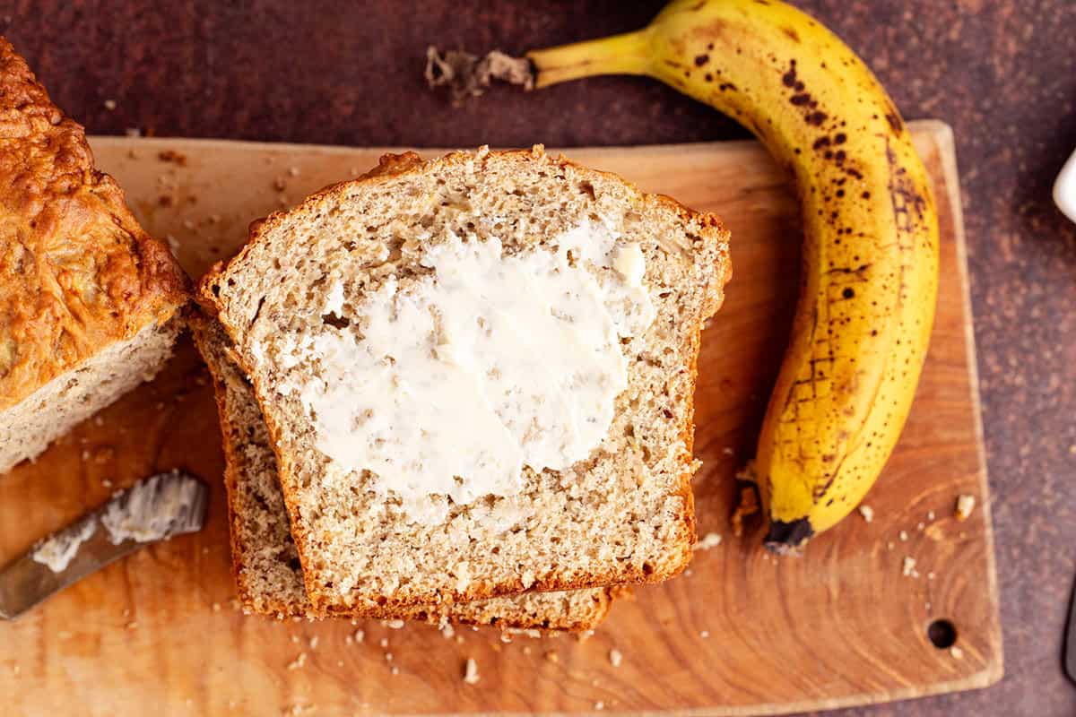 4 Ingredient Banana Bread on a cutting board with butter on it and a banana beside it