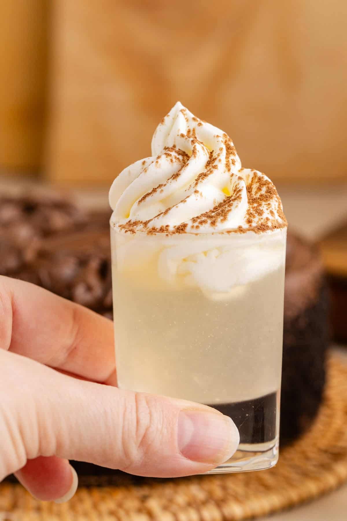 Chocolate Cake Shot in Shot Glass Garnished with Whipped Cream and Cocoa Powder with Chocolate Cake in Background