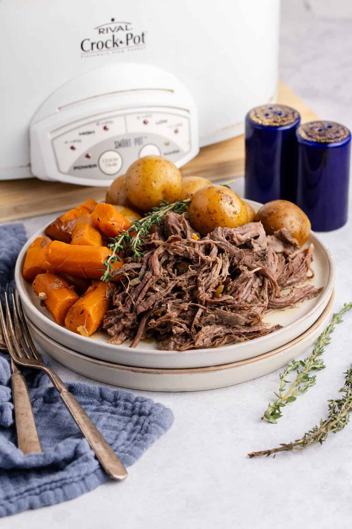 Pot Roast Slow Cooker Recipe all shredded on plate with carrots and potatoes and a slow cooker in the back