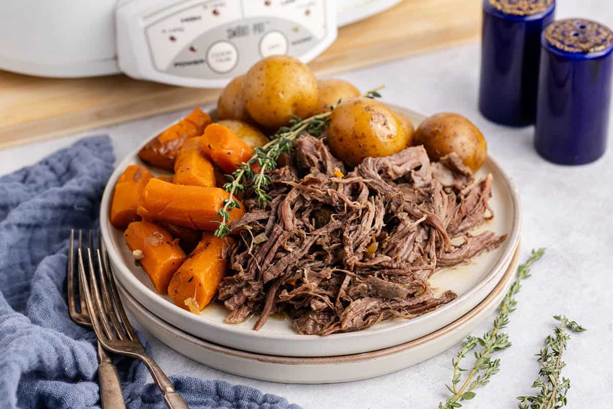 Pot Roast Slow Cooker Recipe all shredded on plate with carrots and potatoes and a slow cooker in the back