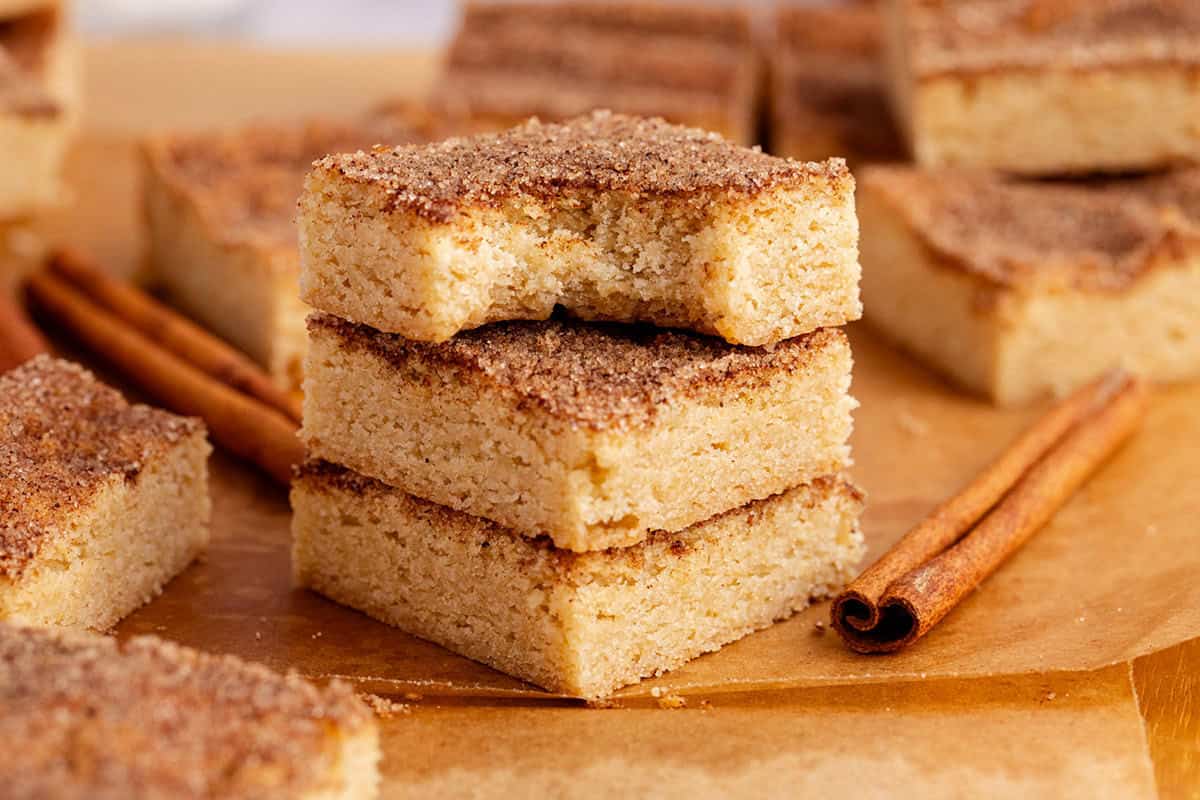 Snickerdoodle Cookie Bars stacked with a bite and cinnamon sticks beside