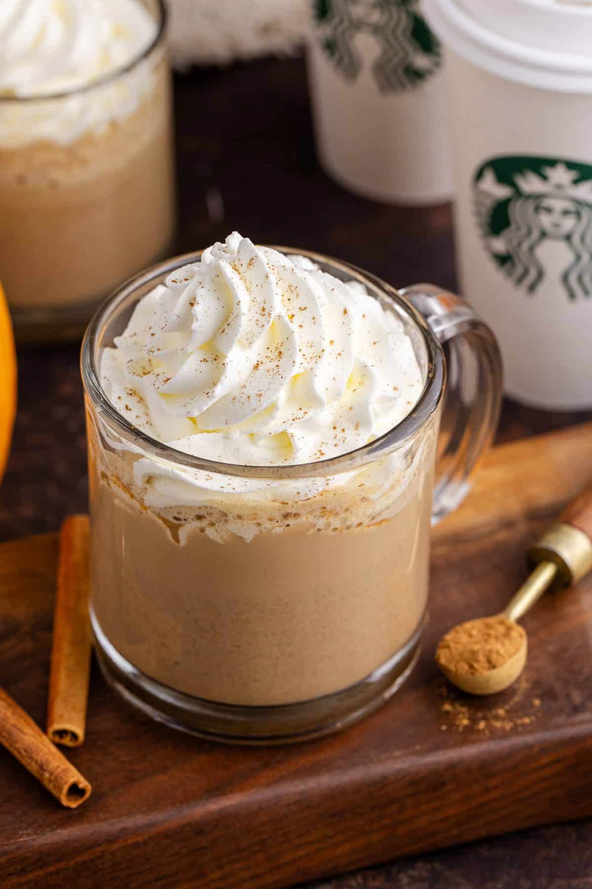 Starbucks Pumpkin Spice Latte Served in a Clear Mug with Whipped Cream and Dusting of Ground Cinnamon with Starbucks Cups in Background