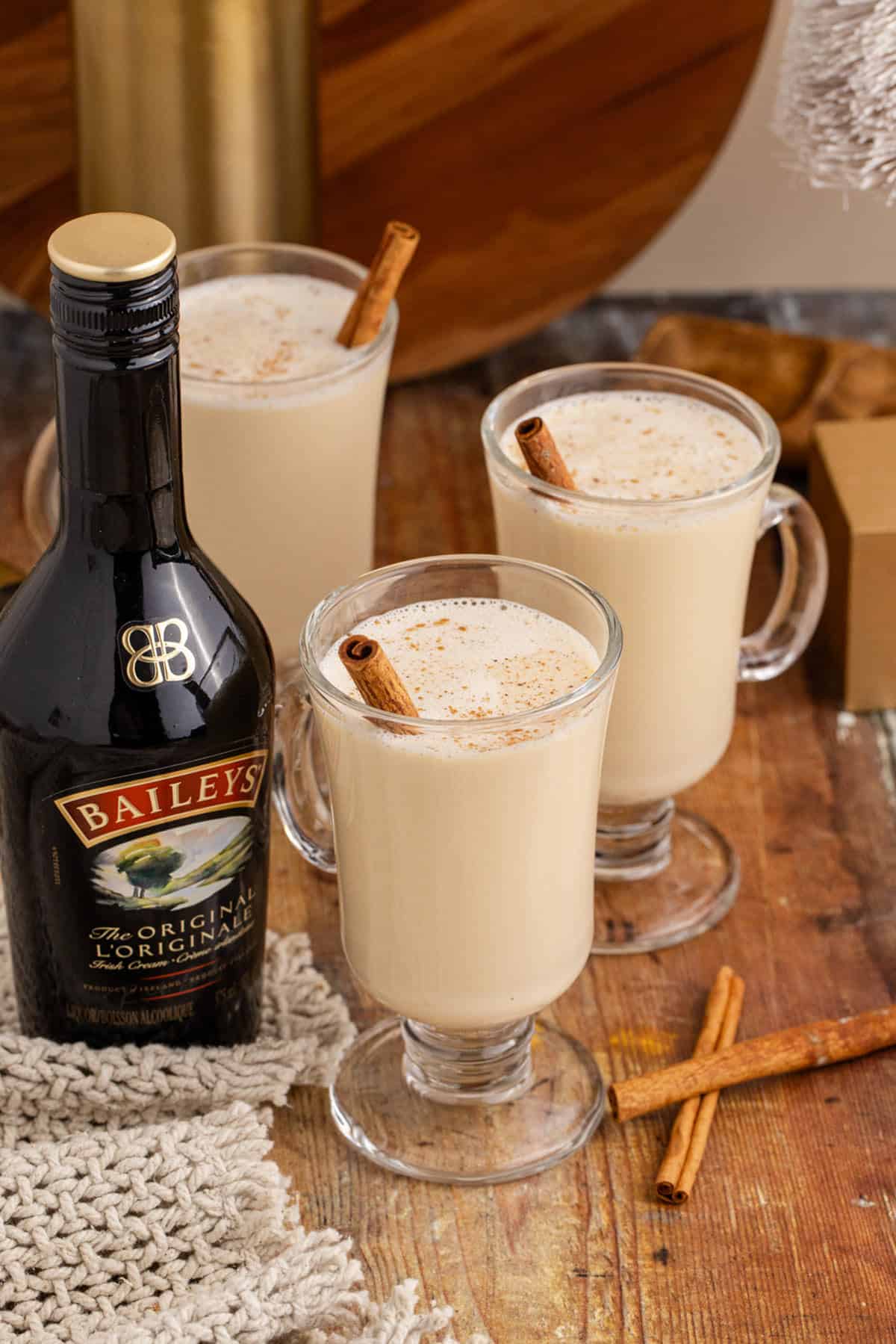 Baileys and Eggnog Served in Tall Glass Mugs with Cinnamon Stick Garnish and Dusting of Ground Cinnamon and Baileys Bottle Beside