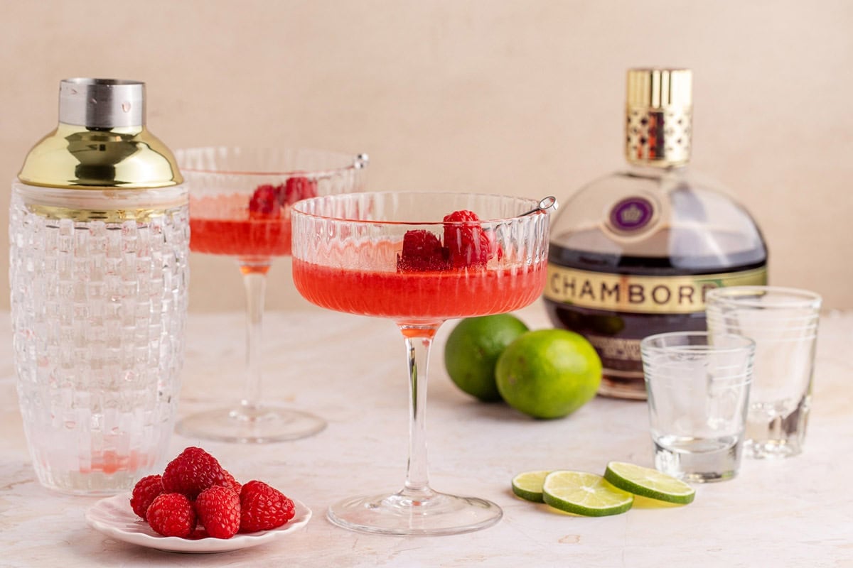 Raspberry Martinis on a counter with limes and a cocktail shaker in the back