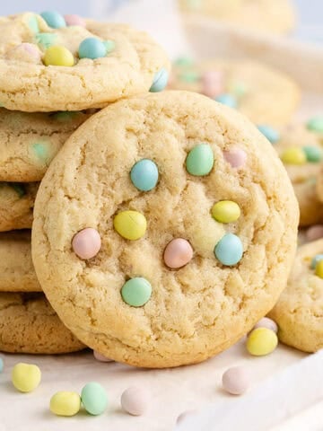 Cadbury Mini Egg Cookies stacked on a baking sheet with mini eggs all around
