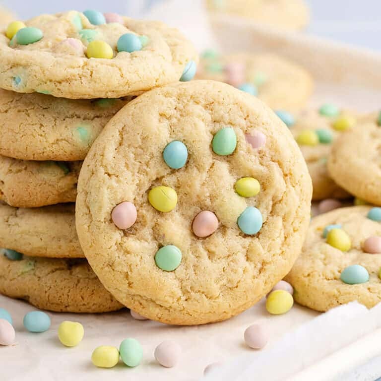 Cadbury Mini Egg Cookies stacked on a baking sheet with mini eggs all around
