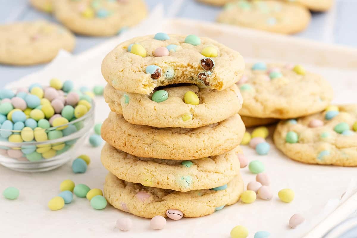 Cadbury Mini Egg Cookies stacked on a baking sheet with a bite taken and mini eggs all around