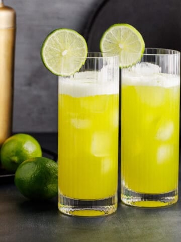 June Bug Cocktails on a counter with lime garnish and bottles in the back