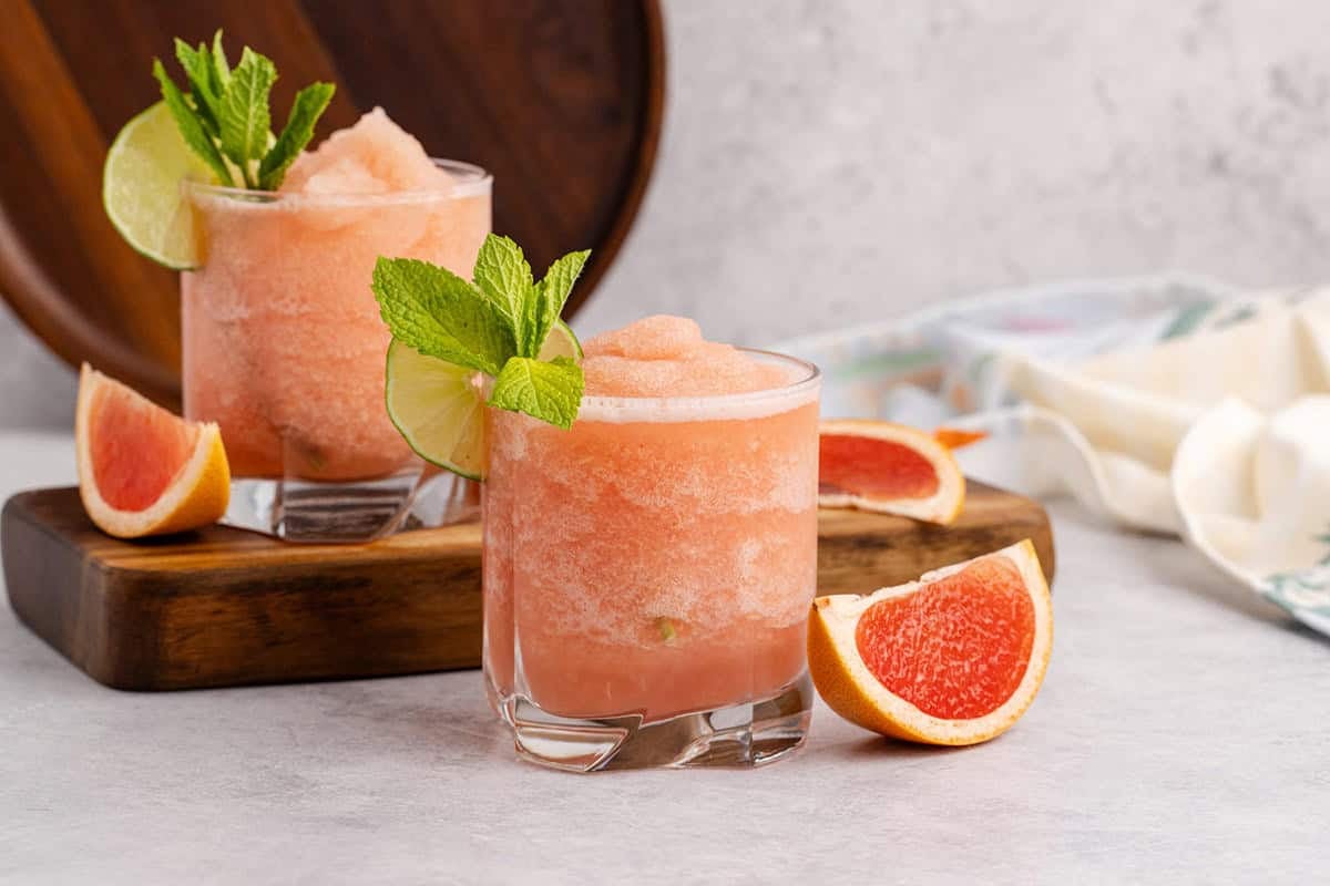 Non alcoholic frozen paloma served in a glass and garnished with a lime round and fresh mint with slices of grapefruit beside and second drink behind