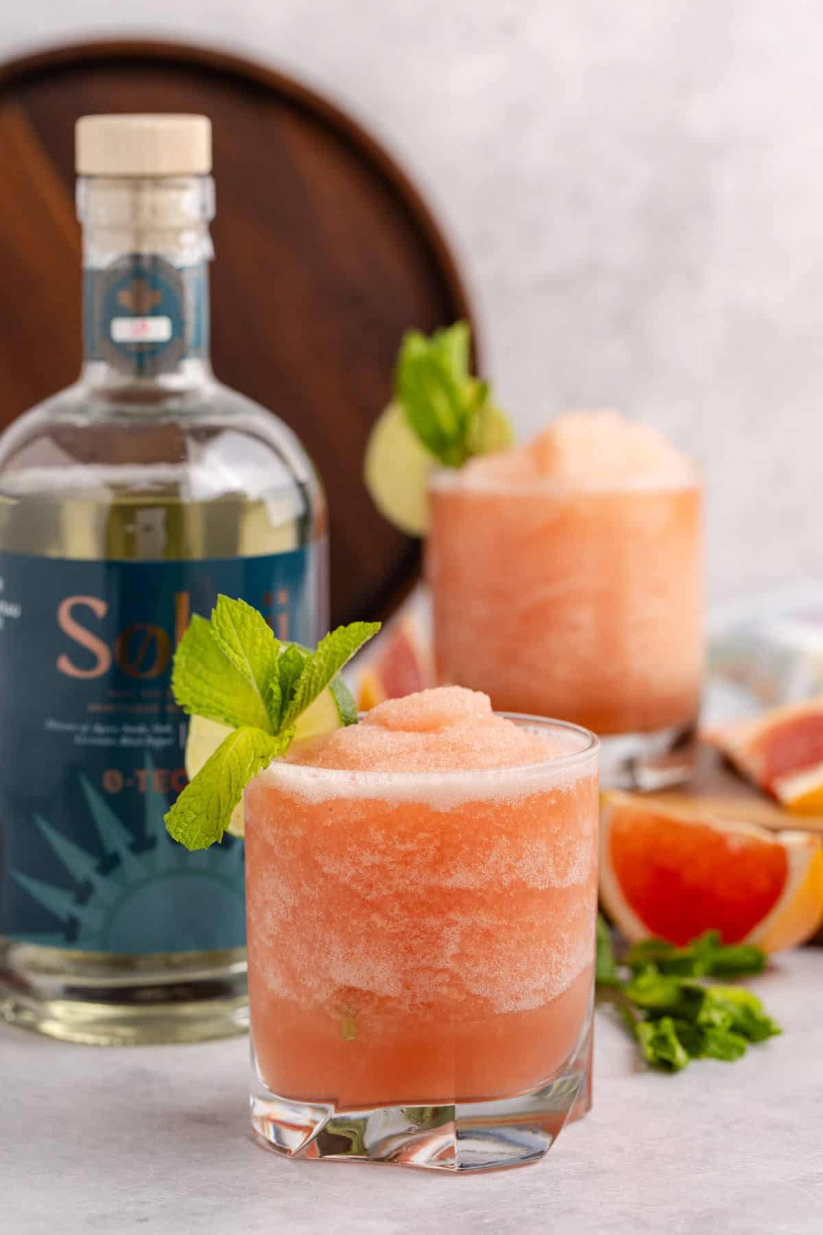 Non alcoholic frozen paloma served in a glass and garnished with a lime round and fresh mint with slices of grapefruit beside and bottle of non alcoholic tequila