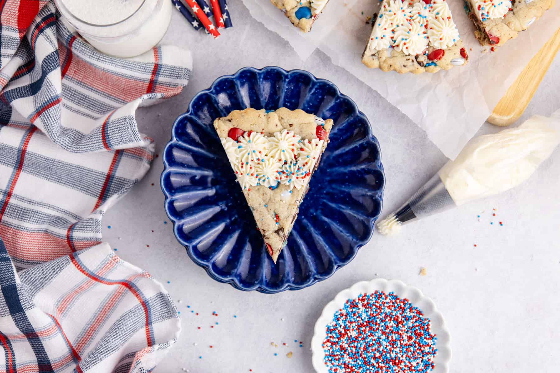 Red White and Blue Cookie Cake from above with a slice on a plate, a glass of milk and sprinkles near