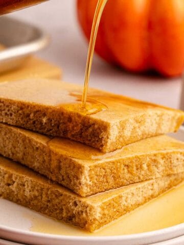 Pumpkin Spice Sheet Pan Pancakes stacked on a plate with maple syrup pouring
