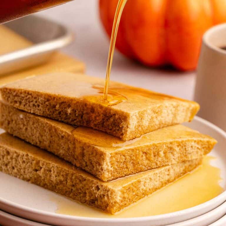 Pumpkin Spice Sheet Pan Pancakes stacked on a plate with maple syrup pouring