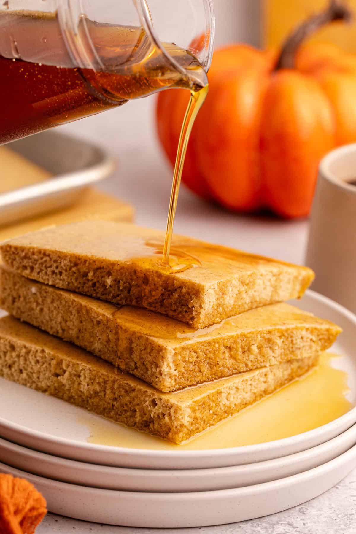 Pumpkin Spice Sheet Pan Pancakes stacked on a plate with maple syrup pouring
