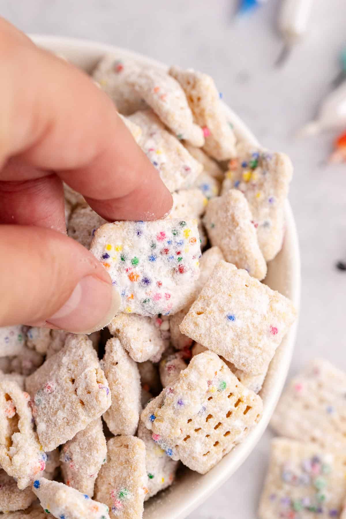 Cake Batter Puppy Chow in a bowl close up with a hand grabbing some